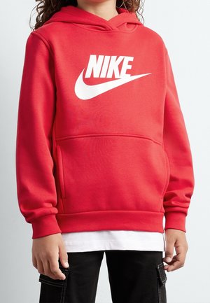 Lapsi, jolla on punainen Nike-huppari valkoisella logolla, mustat taskulliset housut ja hevauksen alta näkyvä valkoinen paita.