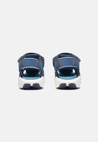 Sininen sandaali puoliksi avoimella muotoilulla, jossa on säädettävät hihnat, valkoinen pohja ja oranssit yksityiskohdat päällisessä. Teksturoitu pohja näkyvissä.