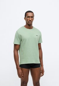 Πράσινο μπλουζάκι με υφή, κοντά μανίκια, στρογγυλή λαιμόκοψη και μαύρο λογότυπο στο στήθος, φορεμένο με μαύρο σορτς. Απλό φόντο.