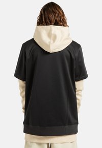 Felpa sportiva nera con cappuccio beige, maniche corte e finitura texturizzata; indossata sopra una maglietta a maniche lunghe beige.