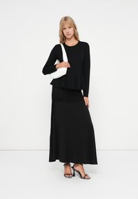 Schwarzes Langarmshirt mit einem ausgestellten Saum, kombiniert mit einem schwarzen, gerafften Maxiröcke. Eine weiße Handtasche und schwarze, schnörkellose Sandalen mit Absatz ergänzen den Look.