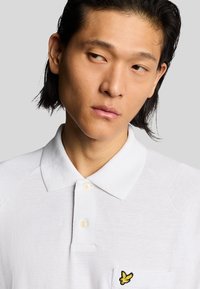 Hvid polo t-shirt med tekstureret stof, to knapper og et gult broderet logo på venstre bryst. Den har en klassisk krave og raglanærmer.