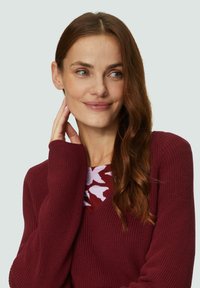 Bordeaux gebreide trui met een V-hals, ribbeltextuur en langere mouwen. Onder deze trui is een gebloemd topje in rood en wit zichtbaar.