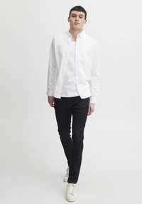 Camisa blanca de manga larga con botones sobre una camiseta blanca, combinada con vaqueros oscuros y zapatillas beiges, llevada por un modelo masculino de pie.