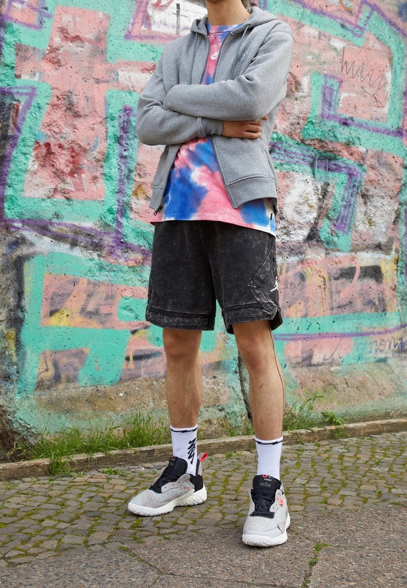 Personne debout, bras croisés, portant un sweat à capuche gris, un t-shirt tie-dye, un short noir, des chaussettes blanches et des baskets noir et blanc devant un mur de graffitis.
