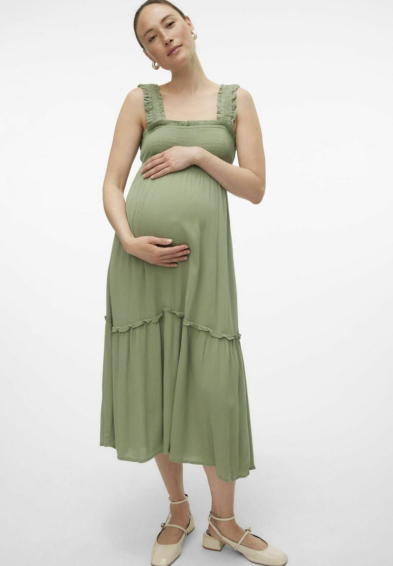 Vestido de maternidad en verde claro, con tirantes con volantes, un corsé ajustado y una falda escalonada. Combinado con zapatos de tacón abierto beige.