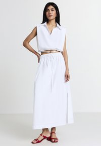 Weiße ärmellose Bluse mit V-Ausschnitt, kombiniert mit einem weißen Maxirock mit elastischem Bund und seitlichem Schlitz. Rote Absatzsandalen.