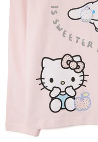 Lichtroze katoenen T-shirt met een afbeelding van een witte kat met een strik, die zijn pootjes vasthoudt, en een appel met sprankelende accenten.