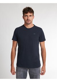 Maglietta in cotone blu navy con maniche corte, colletto rotondo e un piccolo logo ricamato sul petto. Indossata con jeans grigi.