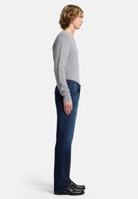 Maglione grigio, jeans in denim blu, vista laterale. Il maglione ha maniche lunghe, i jeans sono a sigaretta, le scarpe sono nere con una finitura lucida.
