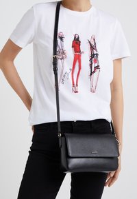 Black crossbody bag with a textured finish, gold-tone hardware, and a flap closure, worn with a white T-shirt featuring fashion illustrations.