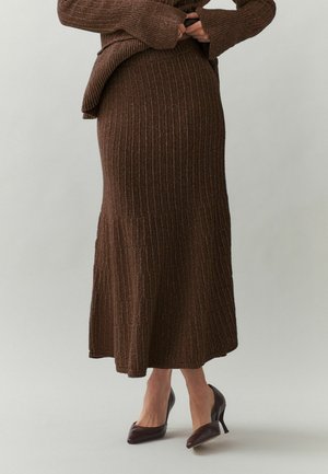 Naine, kes kannab pruunist soonikkoes midi seelikut ja sobivat kampsunit, tumedate pruunide tipptallaga kontsakingadega ühevärvilisel taustal.