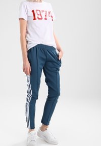 Vit t-shirt med röd "1974" tryck, parat med turkosa joggingbyxor med vita sidostripes och elastisk midja, accentuerad med vita sneakers.