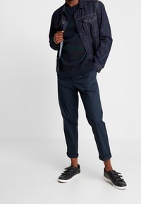 Giacca di denim blu scuro; maglione navy con righe orizzontali; pantaloni navy slim-fit; sneakers nere con tre strap; outfit casual.