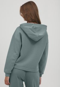 Felpa leggera blu-grigia con una vestibilità rilassata, dotata di cappuccio e polsini a coste. Il tessuto appare morbido con una texture liscia.