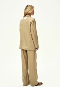 Blondínka stojaca zboku v béžovom obleku so širokými nohavicami a leopardími topánkami na bielom pozadí.