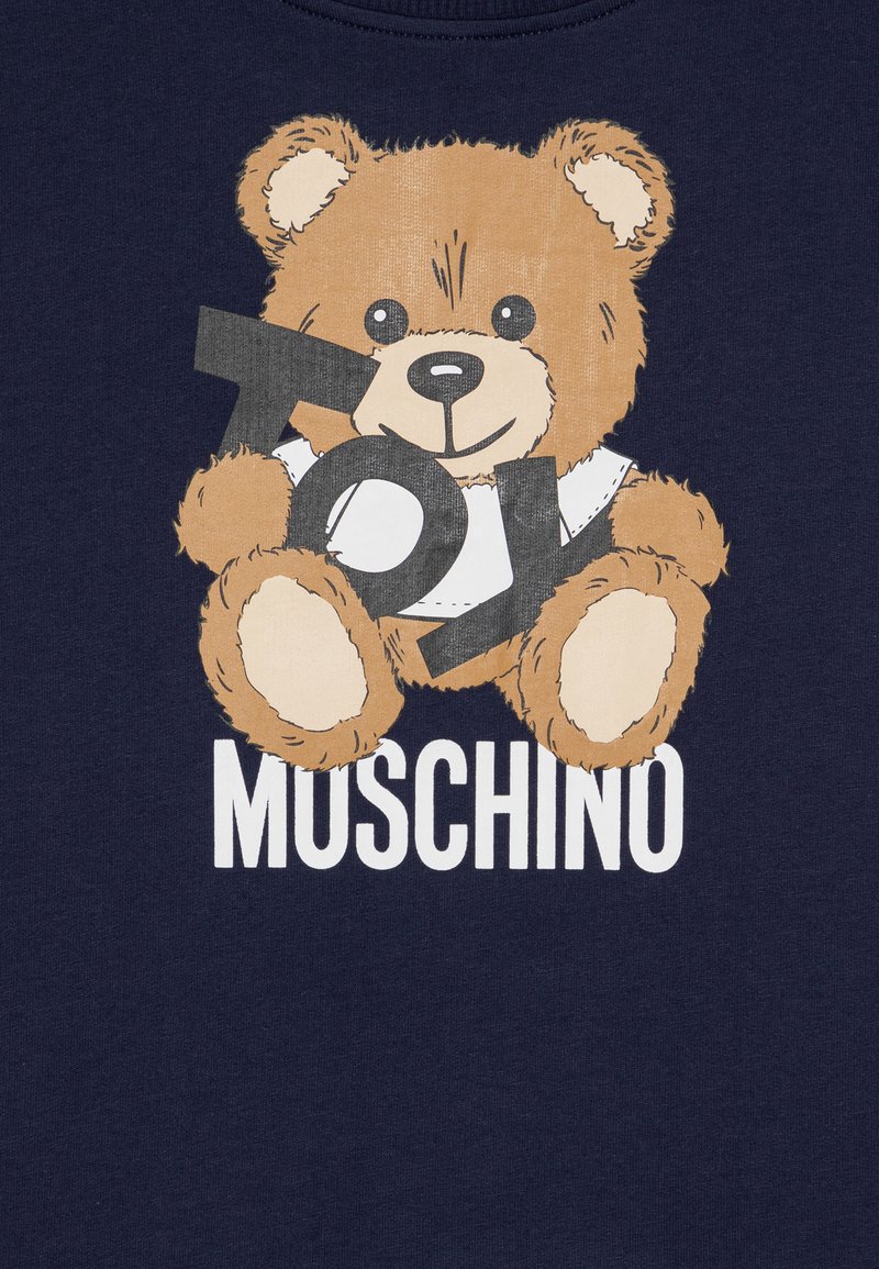 T-shirt navy con grafica di un orso dai peli marroni, che tiene un cartello nero con la scritta "Toy". Il testo in basso recita "MOSCHINO" in lettere bianche in grassetto.