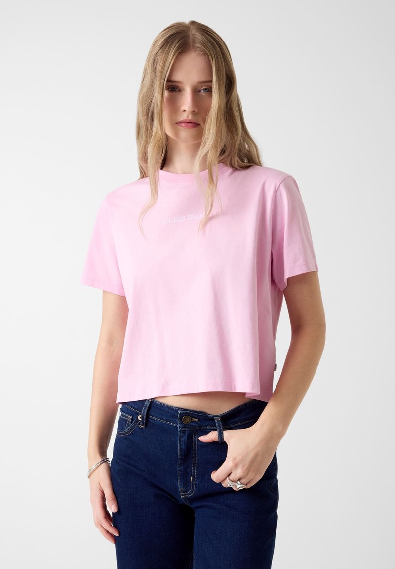 Jasnoróżowy krótki t-shirt z krótkimi rękawami i okrągłym dekoltem, z logo "Guess Jeans" w białym kolorze na przodzie, wykonany z bawełny.