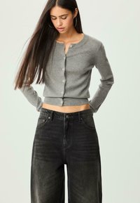 Šedý pletený cardigan s kulatým výstřihem a zapínáním na knoflíky. Nosený s černými džínami s vysokým pasem, zvýrazňující zkrácený střih. Jednoduchý design.