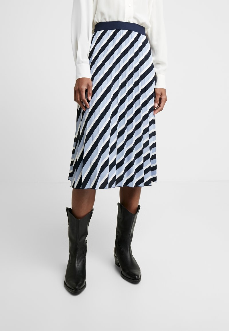 Femme portant une jupe midi plissée à rayures diagonales bleu marine et blanches, associée à des bottes noires mi-mollet et un chemisier blanc.