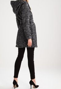 Svart-vit prickig lång jacka med huva, midjeband och fickor på sidan, kombinerad med svarta smala byxor och svarta högklackade skor.