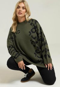 Przesadnie duży oliwkowy sweter z czarnym wzorem zwierzęcym, z prążkowanym dekoltem i mankietami. Połączony z czarnymi spodniami i trampkami.