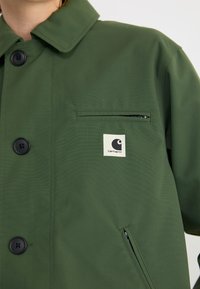 Veste verte en tissu durable avec un col boutonné, une poche poitrine et un patch logo. Elle dispose d'une poche zippée sur le côté et d'une texture mate.