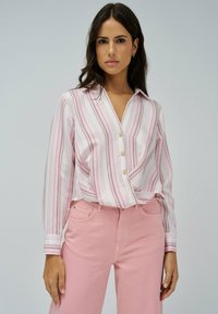 Blusa a rayas en blanco y rosa, con escote en V, cierre de botones y mangas largas. Combinada con pantalones rosa de cintura alta.