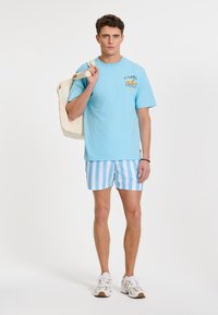 Hellblaues Baumwoll-T-Shirt mit Grafikdruck, kombiniert mit blau-weiß gestreiften Badeshorts. Das Model hält eine cremefarbene Tragetasche.