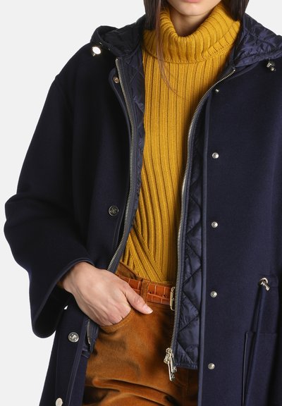 Femme portant un manteau matelassé bleu marine sur un pull à col roulé côtelé jaune moutarde et un pantalon marron avec une ceinture texturée.
