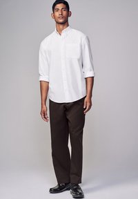 Camicia bianca con maniche arrotolate, dettagli sulla tasca, abbinata a pantaloni marrone scuro e mocassini neri, su uno sfondo neutro.