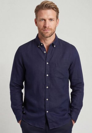 Blonde man met baard die een donkerblauw overhemd met knopen draagt, staat met zijn handen in zijn zakken tegen een effen lichte achtergrond.