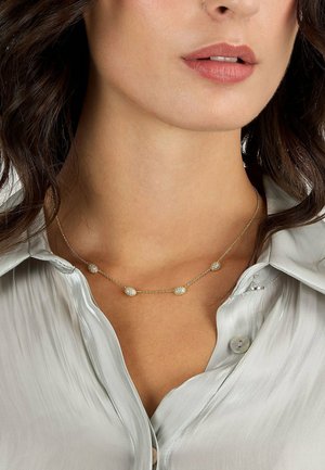 Nahaufnahme einer Frau, die eine goldene Kettenhalskette mit fünf funkelnden ovalen Anhängern über einer hellgrauen Satinbluse trägt.