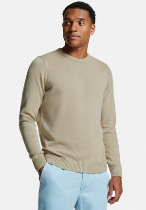 Homme portant un pull Ras-du-cou beige et un pantalon bleu clair, debout avec une main dans la poche, regardant sur le côté sur un fond blanc uni.