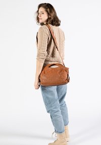 Braune Leder-Umhängetasche mit einem abgerundeten Obergriff, ausgestattet mit einer strukturierten Oberfläche und einem verstellbaren Schulterriemen, vor einem weißen Hintergrund getragen.