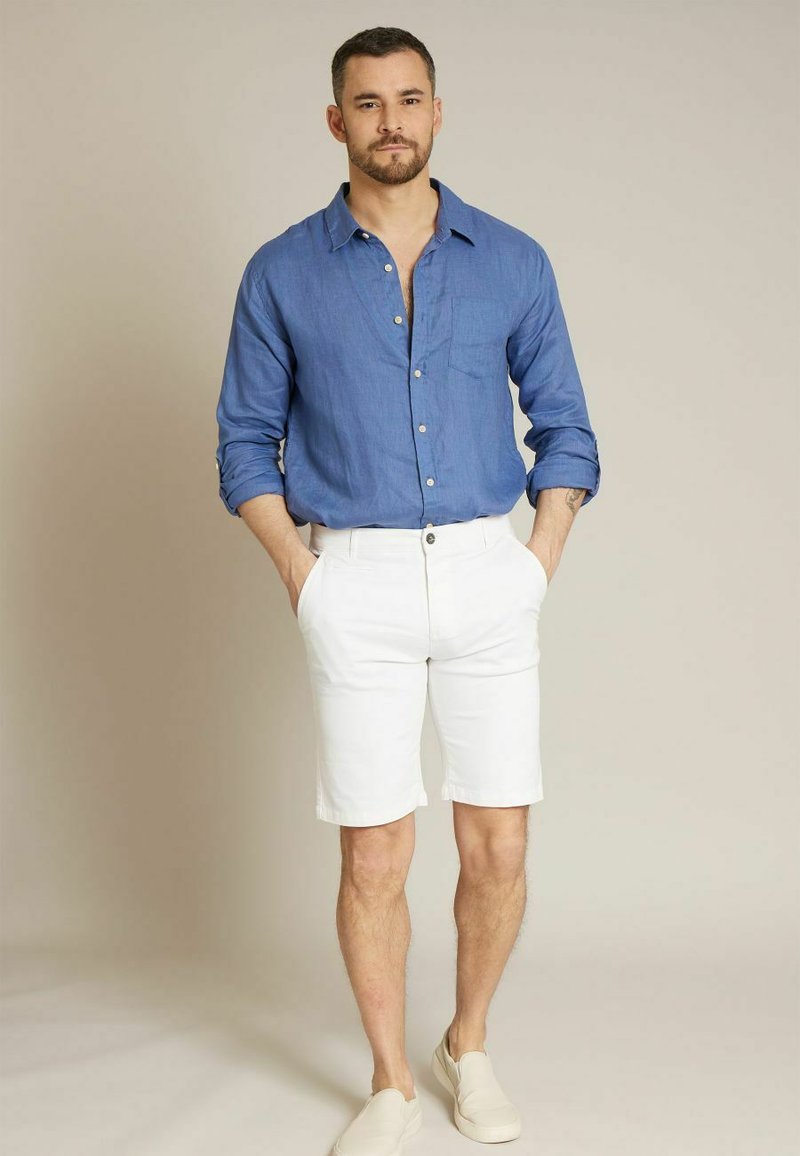 Chemise en lin bleu clair à boutons, shorts blancs et espadrilles beiges. Le mannequin se tient avec les mains dans les poches, montrant une tenue d'été décontractée.