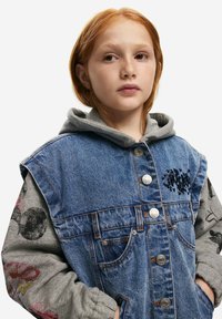 Džínová bunda s kapucí, vyrobená z modré látky a stříbrných patentek. Rukávy mají šedou látku s květinovými vzory a grafikou.