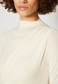 Ljusbeige överdel med hög krage och draperade tygdetaljer. Materialet verkar mjukt och smidigt, med subtila veck vid axeln.