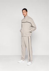 Μπεζ φόρμα με μαύρες ρίγες, κατασκευασμένη από απαλό ύφασμα. Μπουφάν με φερμουάρ και πλαϊνές τσέπες, συνδυασμένο με παντελόνι στο ίδιο σχέδιο και άσπρα παπούτσια.