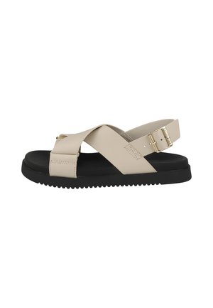 Sandalia beige con tiras cruzadas, hebilla dorada, suela negra y parte inferior texturizada para mejor agarre, mostrada de lado sobre fondo blanco.