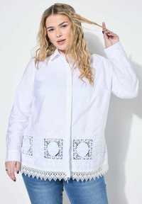 Bílá bavlněná košile s detailními krajkovými akcenty na lemu. Má zapínání na knoflíky a dlouhé rukávy. Hladká textura, klasický design.