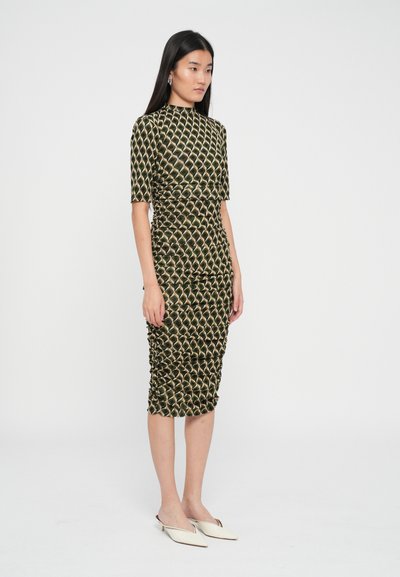 Kobieta stoi w dopasowanej, marszczonej, wzorzystej sukience midi z rękawami do łokci i spiczastych białych klapkach na jednolitym tle.