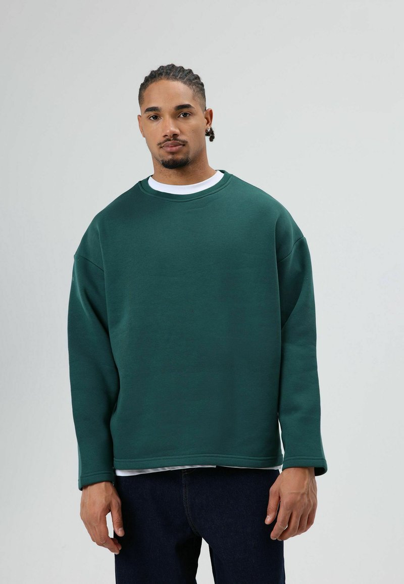 Oversized groene trui met een ronde hals, verlaagde schouders en een gladde textuur, gedragen over een wit T-shirt. Donkerblauwe spijkerbroek.