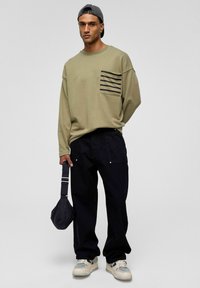 Olijfgroen oversized sweatshirt met een gestreepte zak, gecombineerd met losse zwarte broek en witte sneakers, met een kleine zwarte tas.