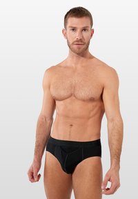 Schwarze Baumwollslips mit einem strukturierten Bund, einer konturierten Fronttasche und einer engen Passform, präsentiert von einer männlichen Figur vor einem neutralen Hintergrund.