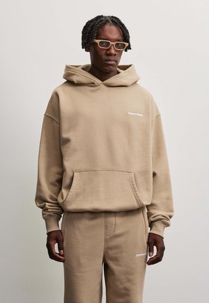 Mand iført beige hættetrøje og matchende bukser med hvide logoer, stående mod en ensfarvet lys baggrund.