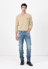 Vaaleanbeige neule, jossa on pieni punainen logo, yhdistettynä vaaleansinihin farkkuihin. Mustat kengät viimeistelevät asun. Yksinkertainen, rento muotoilu.
