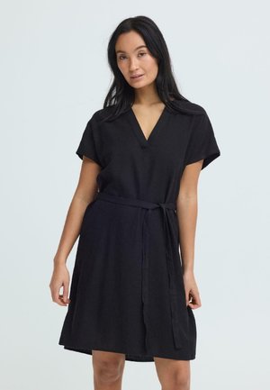 Woman wearing a knee-length black dress with short sleeves, a V-neck, and a tied belt at the waist, standing against a plain background.