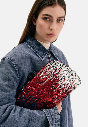 Young woman in denim jacket holding red and silver sequin clutch bag against white background.
