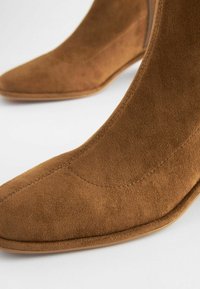 Botine din nubuc maro, cu vârf ascuțit, textură netedă și toc subțire. Prezintă un design uniform, cu detalii de cusătură subtile.